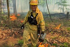 Happy adult in yellow protective gear walks through a forested area with small fires in the background